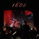 A photograph of a conductor as he conducts a string orchestra in a dark room with a screen and projected abstract, colorful visuals on the upstage wall.