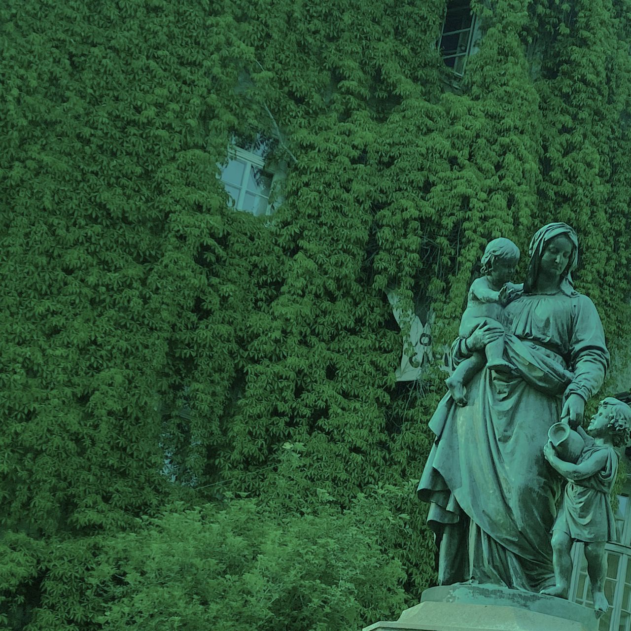 A photograph of a bronze statue of a mother with two children in the foreground, with a building covered in vines and greenery in the background.