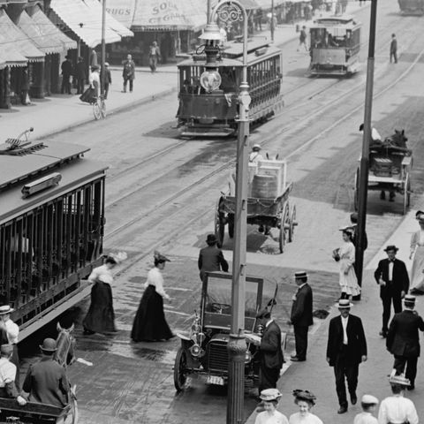 A 19th century monochromatic photograph of an urban landscape with trams, old Ford Model A, horse-drawn wagons, and people visible.