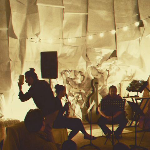 A photograph of a performance with the artist holding and singing into a mask, with the background walls decorated in elaborate hanging sheets of paper.