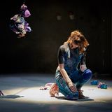 A photograph of the dancer Josseline Black, crouching with her hand placed on her leg, with scattered papers and hanging decorations around her.