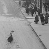 A detail from a monochromatic photograph showing an urban setting with people in the background in 19th century attire.