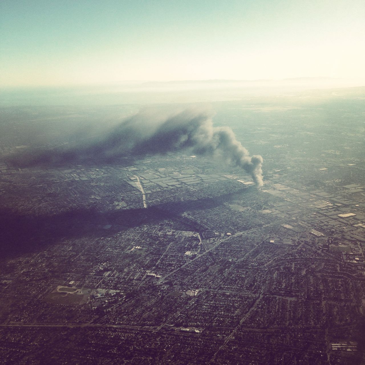 An aerial view of a dense urban area with a fire and smoke cloud casting a large shadow over the city in the distance.