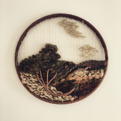 The image is a textile art piece, featuring a circular frame containing a woven landscape. The scene depicts a tree and foliage crafted from various yarns and fibers in shades of green, brown, and beige, giving a textured, three-dimensional effect.