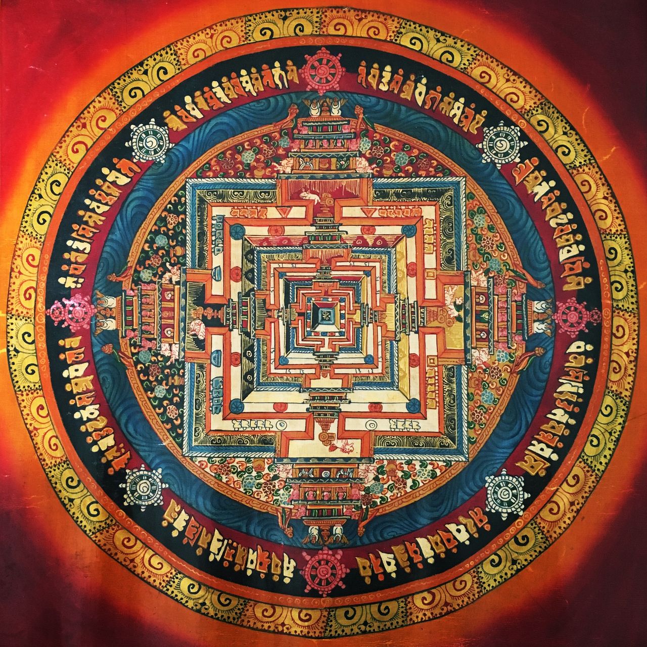 The image is a detailed and vibrant mandala, likely of Tibetan Buddhist origin. It features an intricate, symmetrical design with concentric layers radiating from a central point. The mandala includes elaborate architectural motifs resembling temples, surrounded by various symbols, deities, and patterns.